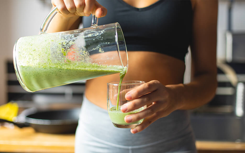 A fit-looking woman in black sports bra and gray leggings pours green juice from a glass pitcher into a glass she's holding; easy smoothie recipes